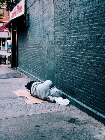 A homeless man lies alone on the street next to a bare brick wall.