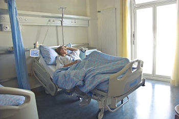 A woman lays in a hospital bed with her hand on her head, deep in thought.