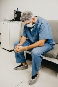 A male physician wearing scrubs, hairnet, and mask seated with hands clasped and looking down.