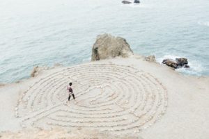 A person walking into the centre of a circular pattern etched out into the sand on a beach.