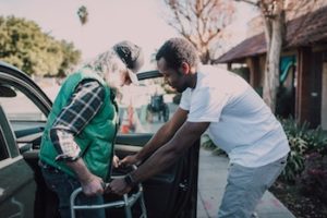 A volunteer helping an older man with a walker into a car.