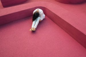 Woman sitting on red carpeted floor in a corner, bent over with her face in her knees.