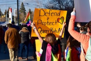 A child walking amongst people in a Pride parade, holding up a sign that reads "Defend & Protect #QueerKids".