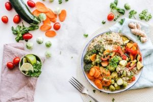 Bowl of quinoa and vegetables laid down on a white linen cloth, alongside a metal fork and more vegetables strewn about.