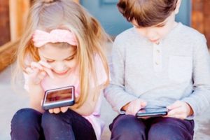Two children sitting side-by-side the front step in front of a door using smartphones.