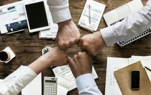 Four hands in a group fist-bump over a work table with paper, coffee, and a calculator strewn across it.