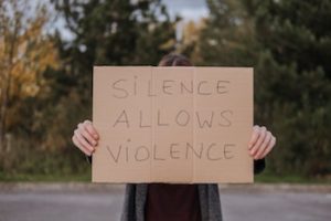 A cardboard sign held up in front of a person reading "Silence Allows Violence".