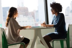 Two women talking at a sunlit table indoors.