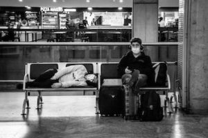 Black and white photo of a man sitting in an airport with a mask on and a woman laying down beside him