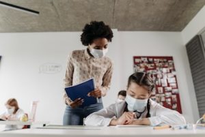 Female teacher wearing a mask covering her mouth and nose, in a classroom of young students also wearing masks, overlooking a young female student's work at her desk.