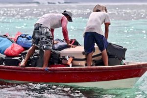 Colour photo of stack of luggage on boat in the water with two people moving luggages