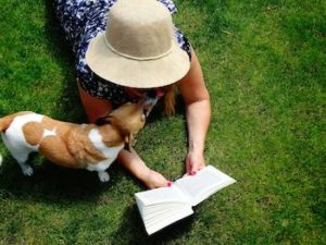 person in white hat sits on the grass and reads to their dog while the dog licks their the humans face