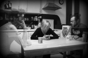 Black and white photo of a woman sitting at a table with ghost-like people beside her