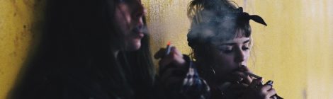 Two women sitting against a dirty wall smoke cigarettes
