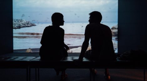 Two people sit talking in the dark, facing a window to a dreary landscape