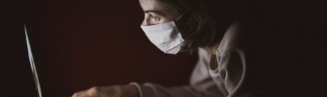 A woman in a surgical mask types on a laptop computer in a dark room.