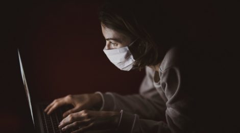 A woman in a surgical mask types on a laptop computer in a dark room.