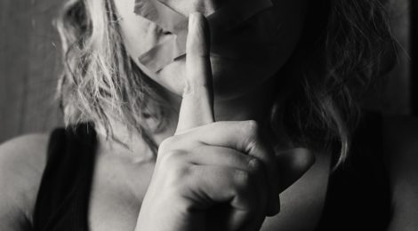 A woman holds a finger up to her tape covered mouth to indicate silence.