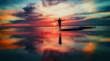 Person standing with their hands open on a beach with the water reflecting a colourful sky.