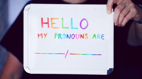 Someone hold a small whiteboard, with the following written in mulit-colour marker: "HELLO / MY PRONOUNS ARE _____/_____".