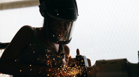Close-up silhouette of a woman wearing a face shield conducting metal work, with sparks in the foreground.