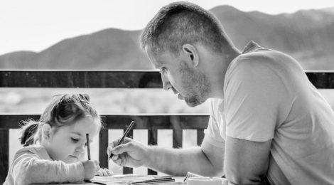 Black-and-white photo of a father and daughter sitting at a table next to the water, colouring or drawing.